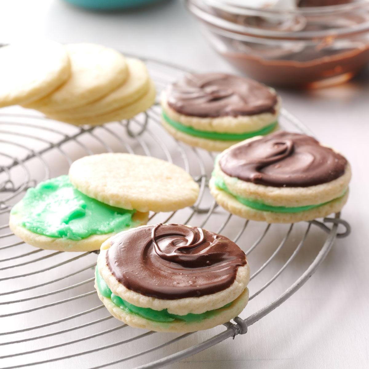 Cookies with green and chocolate frosting rest on a metal rack, while a glass bowl with chocolate sits in the background.
