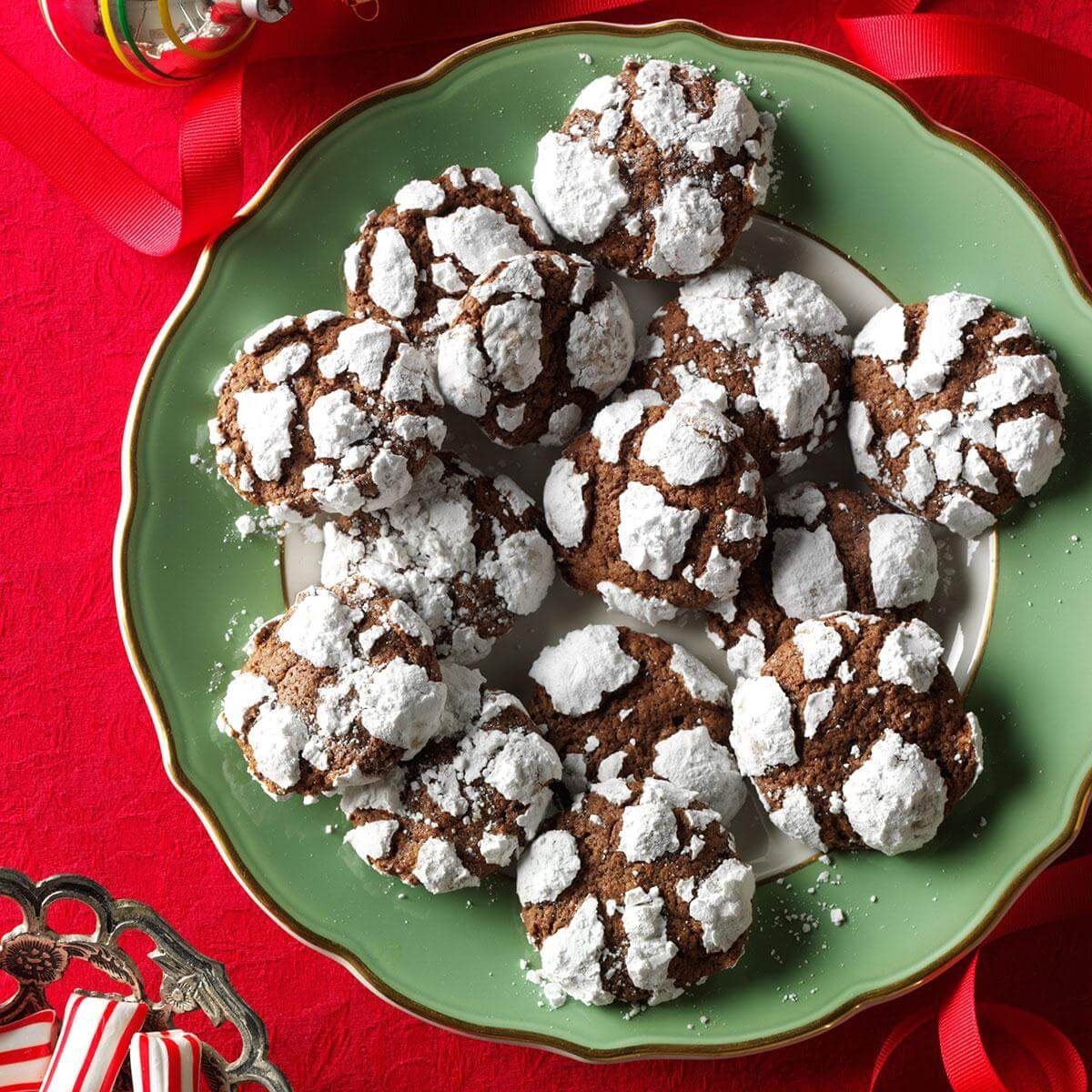 Cookies dusted with powdered sugar rest on a green plate, placed on a red tablecloth with festive decorations nearby.
