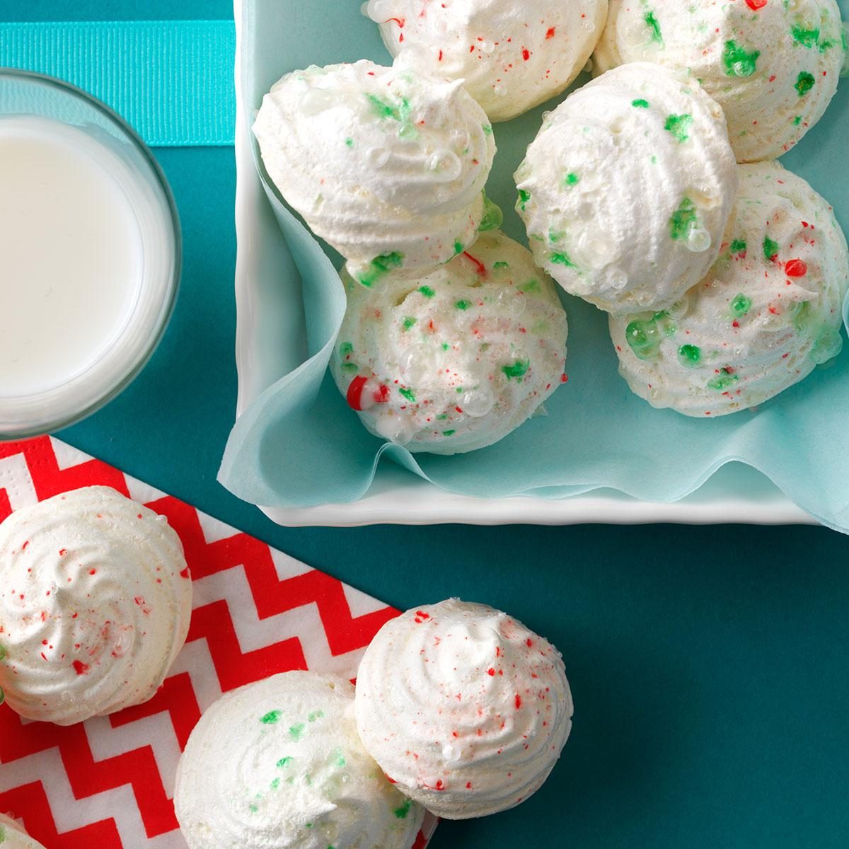 Meringue cookies dotted with red and green sprinkles resting on a teal surface near a glass of milk and red chevron-patterned napkin.