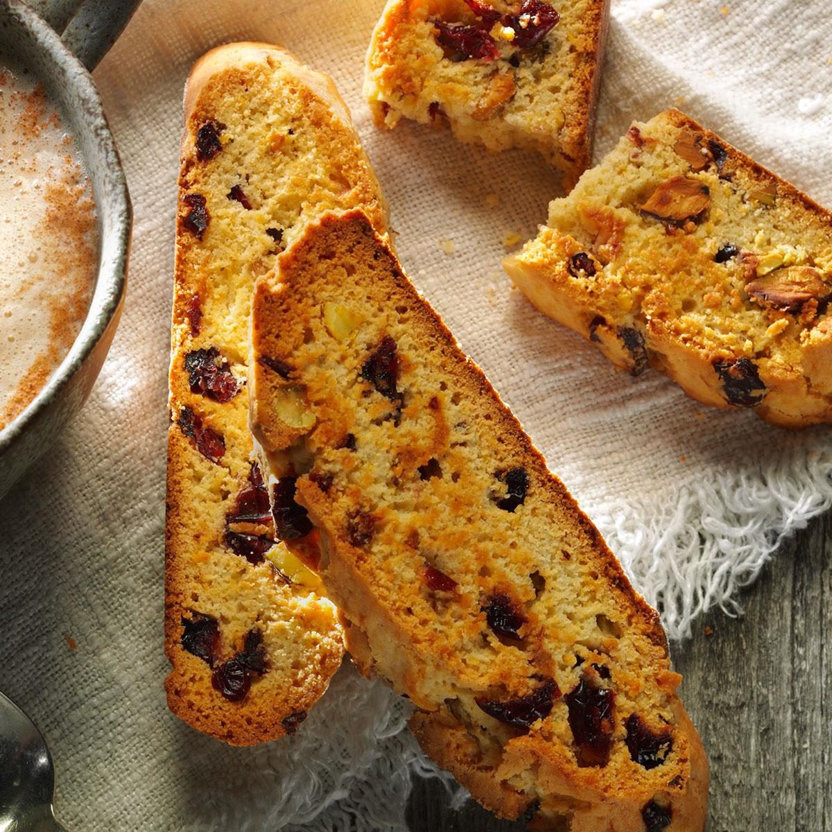 Biscotti resting on cloth, featuring dried fruit and nuts, next to a textured ceramic cup of frothy coffee on a wooden surface.