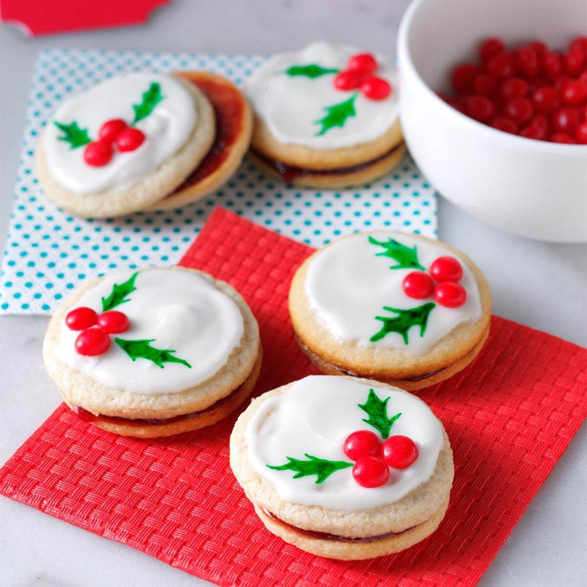 Cookies decorated with white icing and holly designs lie on red and blue checkered napkins, alongside a bowl of red candies.