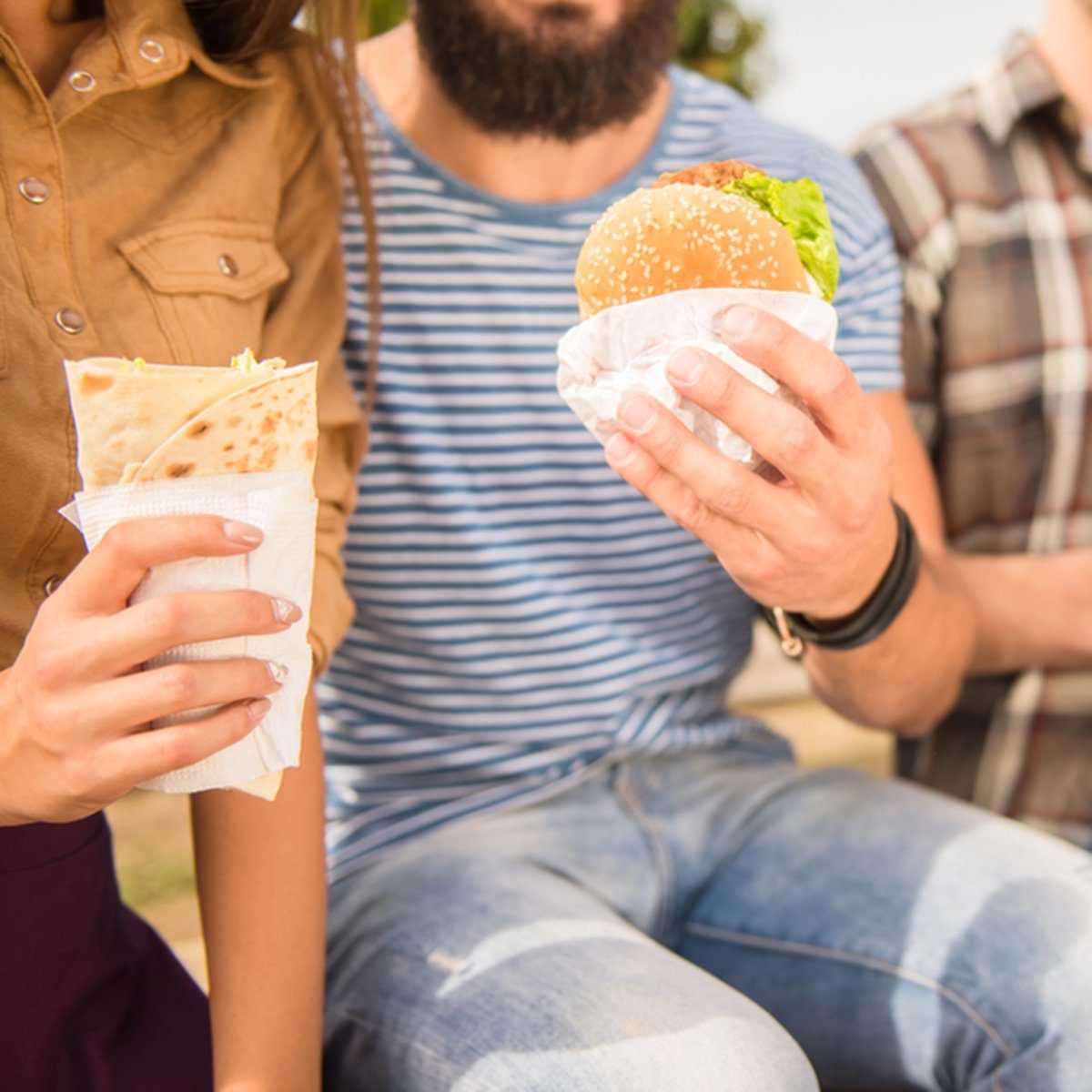 Hands hold a wrap and a burger, seated outdoors.