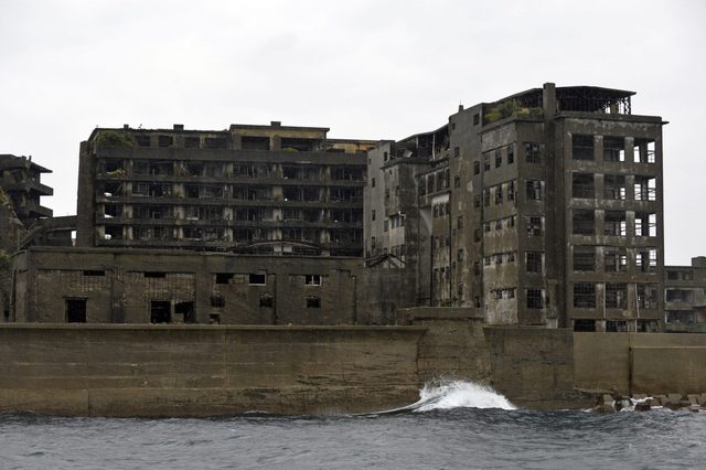 Dilapidated concrete buildings stand abandoned, showing broken windows, in an industrial setting beside a rough sea and overcast sky.