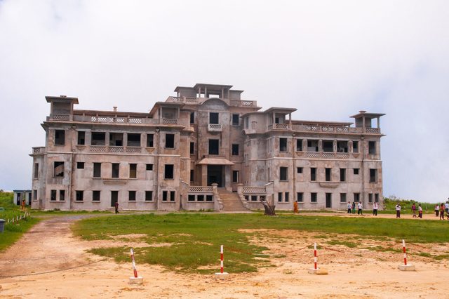 A large, abandoned building sits in an open grassy area with a group of people standing nearby.