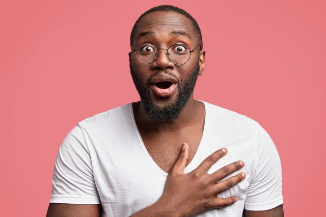 A surprised person with glasses gestures with hands on chest, against a solid pink background.