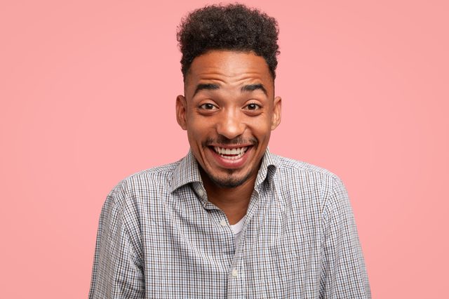 A smiling person faces forward, wearing a checkered shirt against a solid pink background.