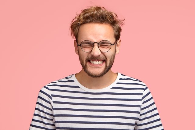 A man with glasses smiles widely against a pink background, wearing a striped shirt.