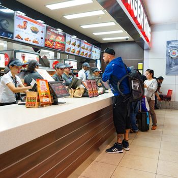 Customers order at a fast-food counter, while employees take orders in a brightly lit restaurant with menu boards displaying food options.