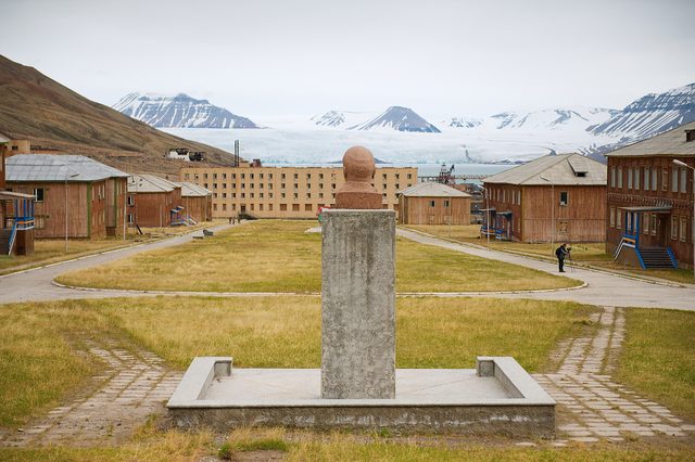Bust sits on a concrete pedestal, overlooking abandoned buildings; snowy mountains and glacier span the distant background under a cloudy sky.