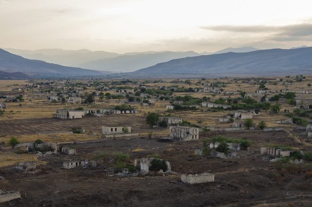 Ruined buildings lie scattered across a desolate, grassy landscape, with distant hazy mountains under an overcast sky.