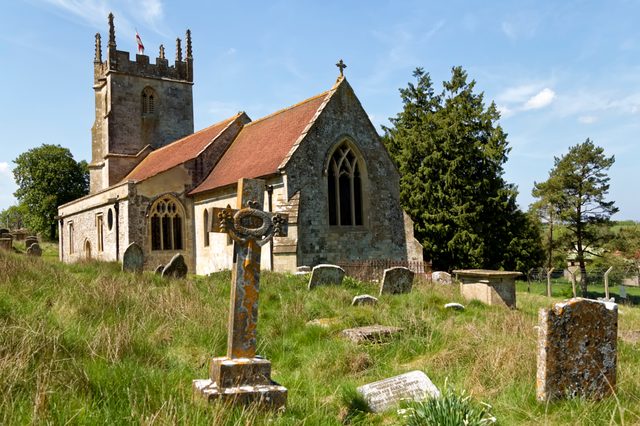 Stone church stands quietly amid an overgrown graveyard, surrounded by trees and grass under a clear blue sky.