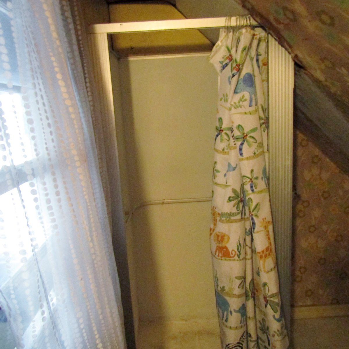 Shower curtain hangs, displaying animals and plants, in a small attic bathroom with slanted walls and patterned wallpaper.