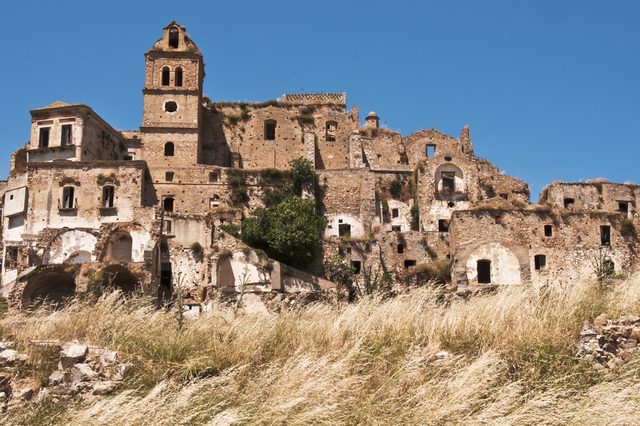 Ancient stone buildings stand abandoned, partially ruined, surrounded by wild grassy fields under a clear blue sky.