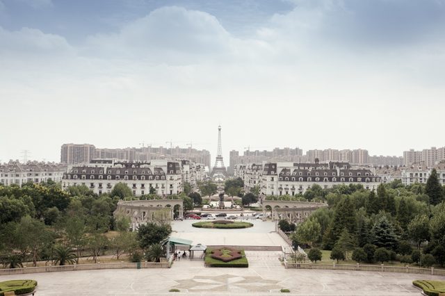Eiffel Tower replica stands amidst symmetrical gardens and European-style buildings, surrounded by tall apartment blocks under a cloudy sky.