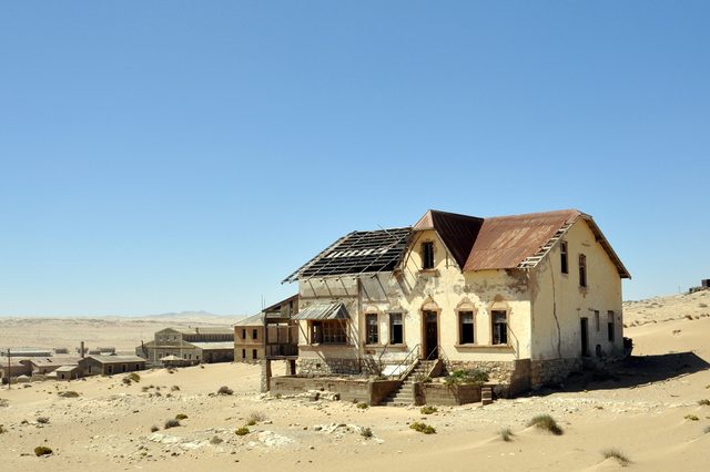Dilapidated house stands abandoned, surrounded by sandy, barren desert with a clear blue sky overhead. Other old buildings visible in the background.