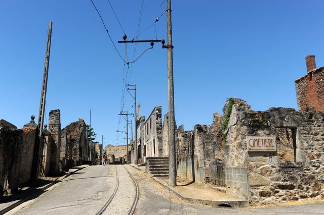 Ruins stand silently under a clear blue sky, electric cables stretch overhead; a sign reads "CimetiÃ¨re".