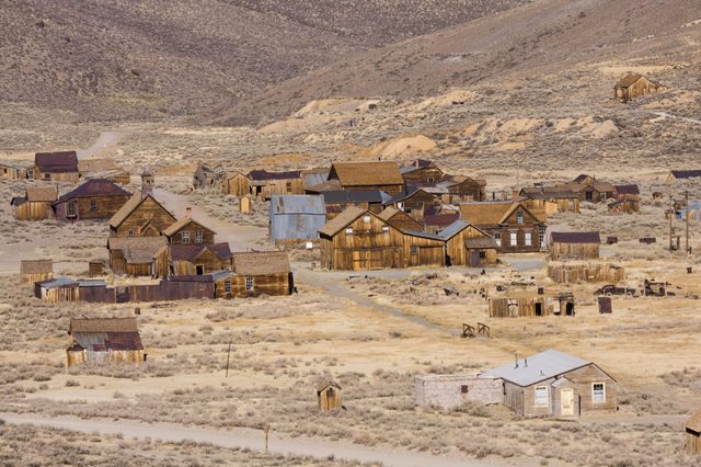 Wooden buildings stand silently in a deserted ghost town surrounded by a barren, hilly landscape with sparse vegetation.