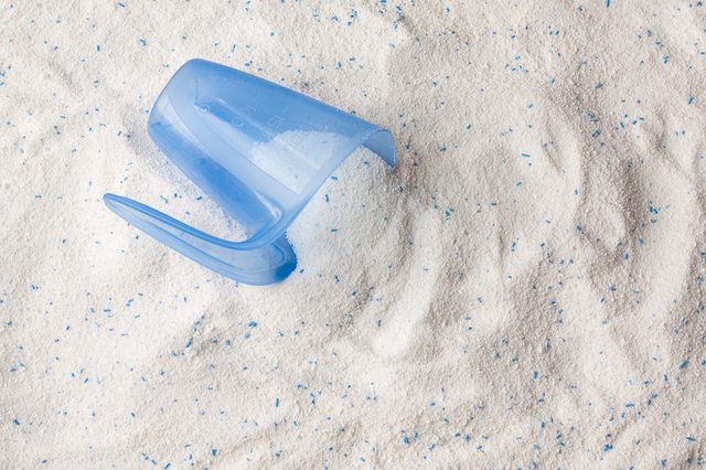 A blue scoop lies partially buried in white granular powder with scattered blue specks, likely detergent, spread across the surface.