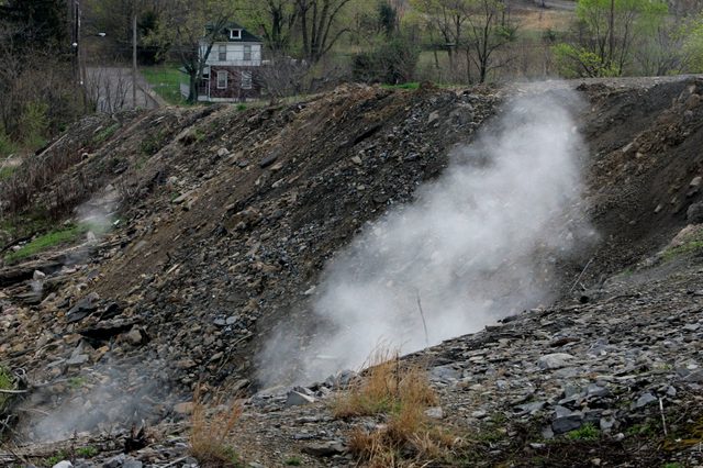 Smoke rises from rocky hillside, surrounded by sparse vegetation, with a small house visible on a distant road.