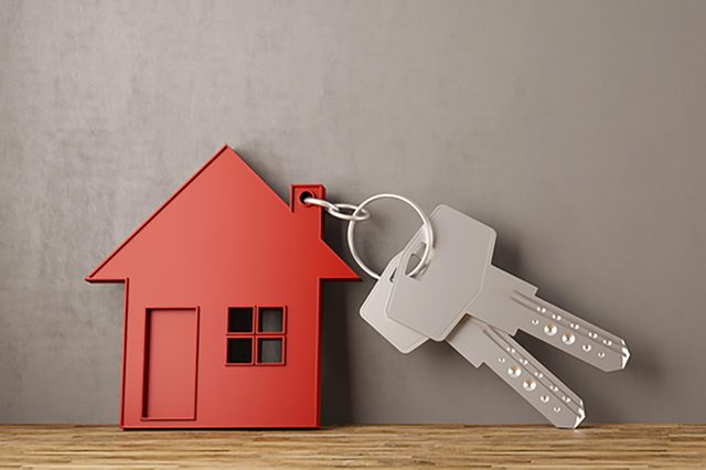 Red house keychain holds keys, lying on a wooden surface against a plain gray wall.