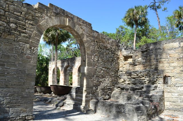 Stone archway stands in ruins, surrounded by tropical trees and fragmented walls under a clear blue sky.