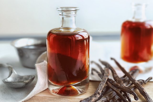 Glass bottle holds brown liquid, standing on a table with vanilla pods and a spoon in soft-focus background.