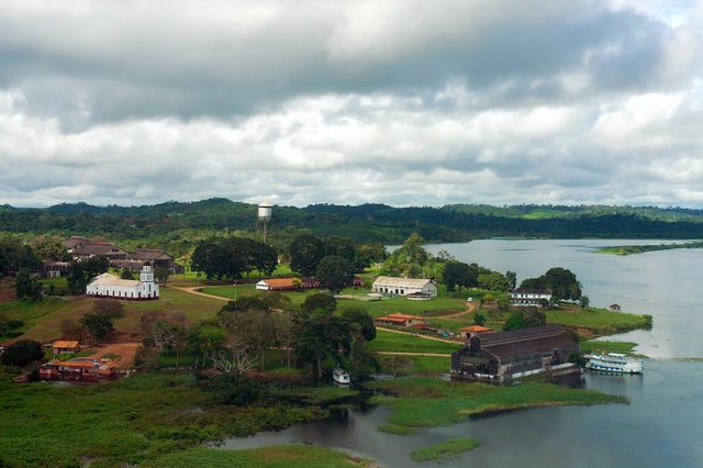 Buildings rest quietly alongside a river, surrounded by lush greenery and distant hills under a cloudy sky.