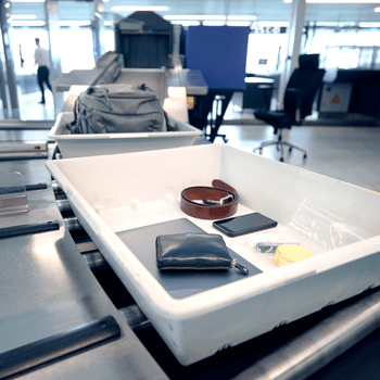 Plastic bin holds personal items on security conveyor. Bags and trays move through airport checkpoint. Office chairs and machines in the background.