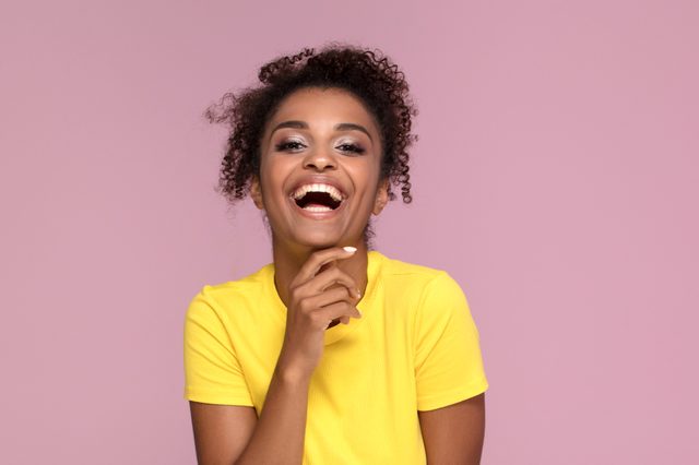 A person in a yellow shirt laughs joyfully against a plain pink background.