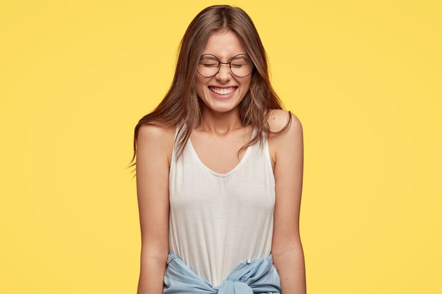A woman smiles broadly while standing calmly, wearing glasses and casual clothes, against a bright yellow background.