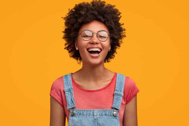 A person wearing glasses and denim overalls is smiling broadly against a solid orange background.