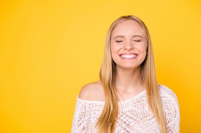 A person, smiling broadly with eyes closed, stands against a vibrant yellow background, wearing a white lace top.