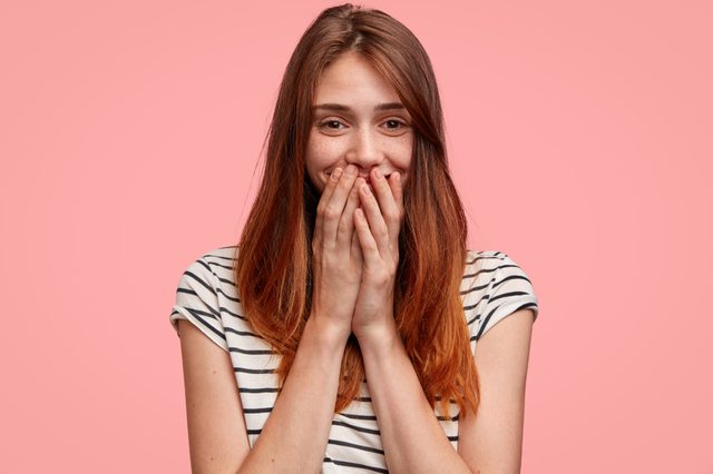 A person covers their mouth playfully, smiling in a striped shirt against a solid pink background.