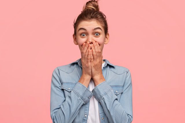 Person covers mouth with both hands, expressing surprise; wearing a denim shirt, in front of a pink background.