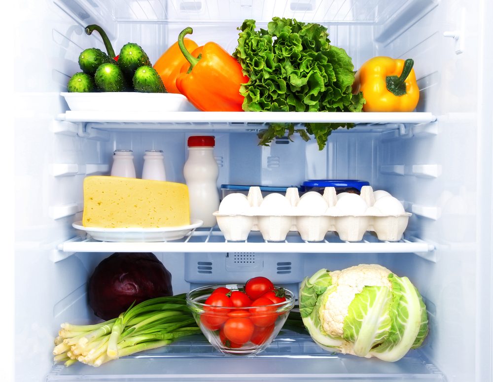 Refrigerator shelves hold fresh vegetables, cheese, eggs, milk, and condiments, neatly arranged in a brightly lit interior.