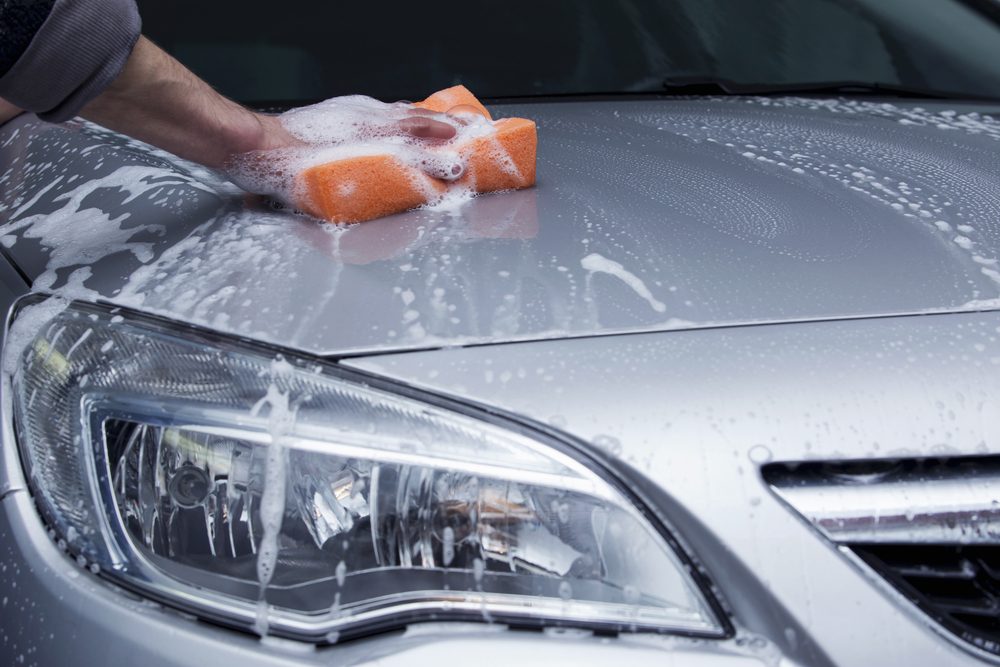 A hand scrubs a car hood with a soapy orange sponge, surrounded by soap suds and water streaks.