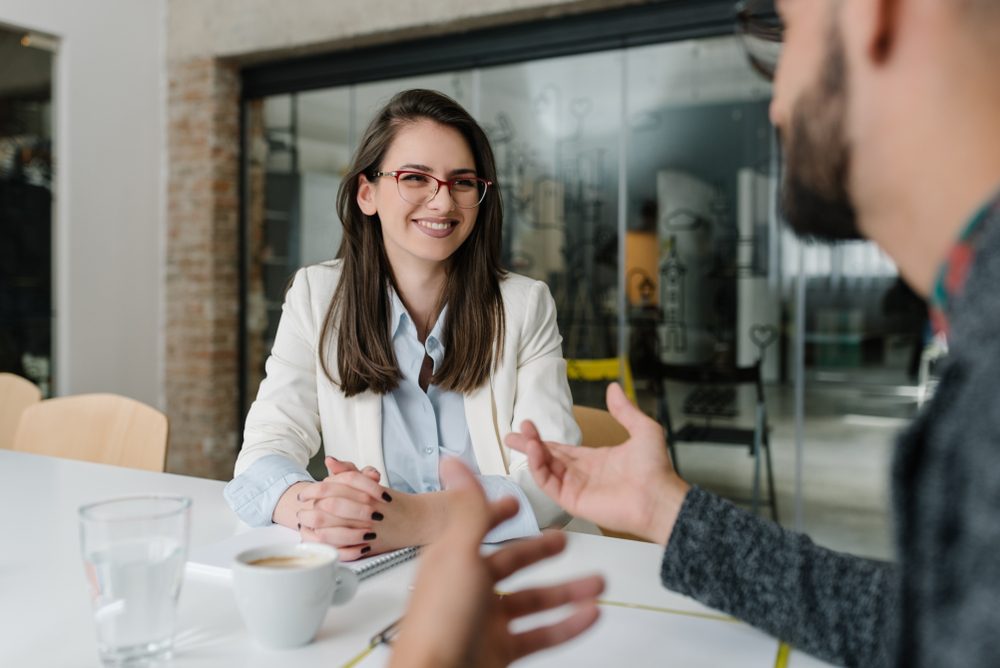 Woman smiles during a conversation at a modern office table with coffee cups and a glass, surrounded by chairs and glass walls.