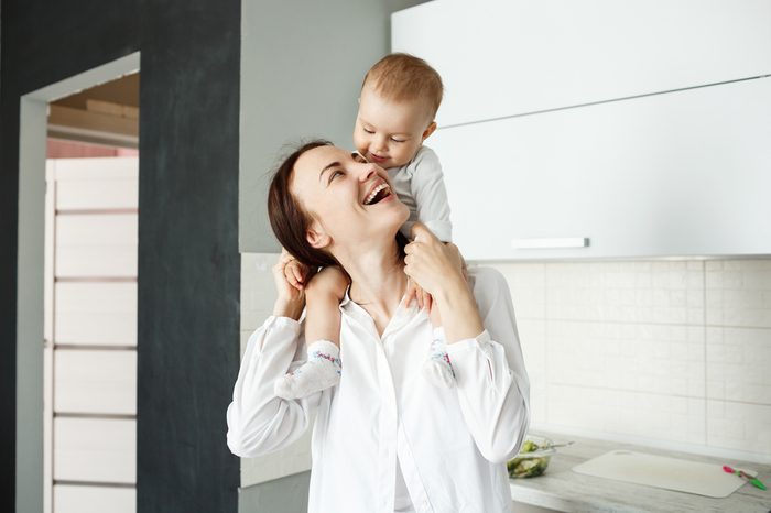 Portrait of happy young mother holding her little precious child on shoulders, looking at him and laughing while baby smiling and looking in mom eyes.