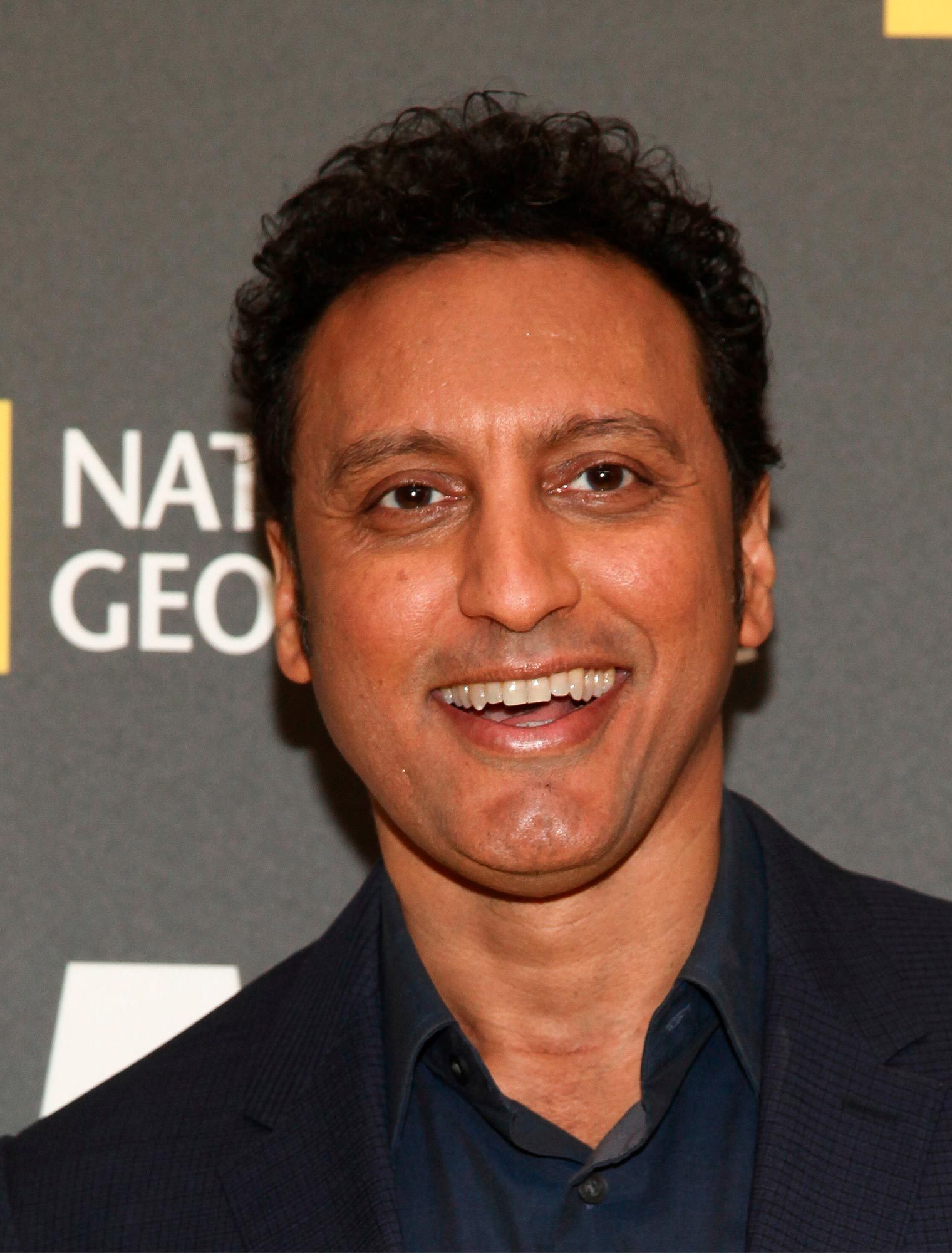 A person smiles widely against a background with "NATIONAL GEOGRAPHIC" text, wearing a dark blazer and shirt.