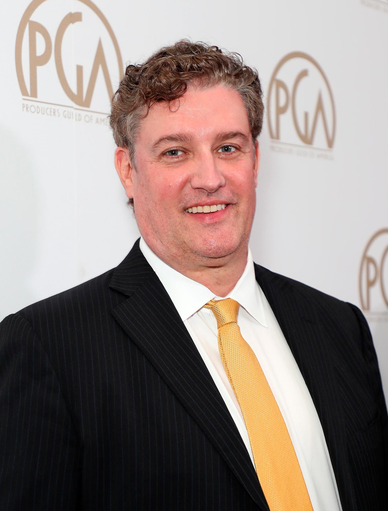A person smiles, wearing a suit with a yellow tie, standing against a backdrop with "Producers Guild of America" logos.