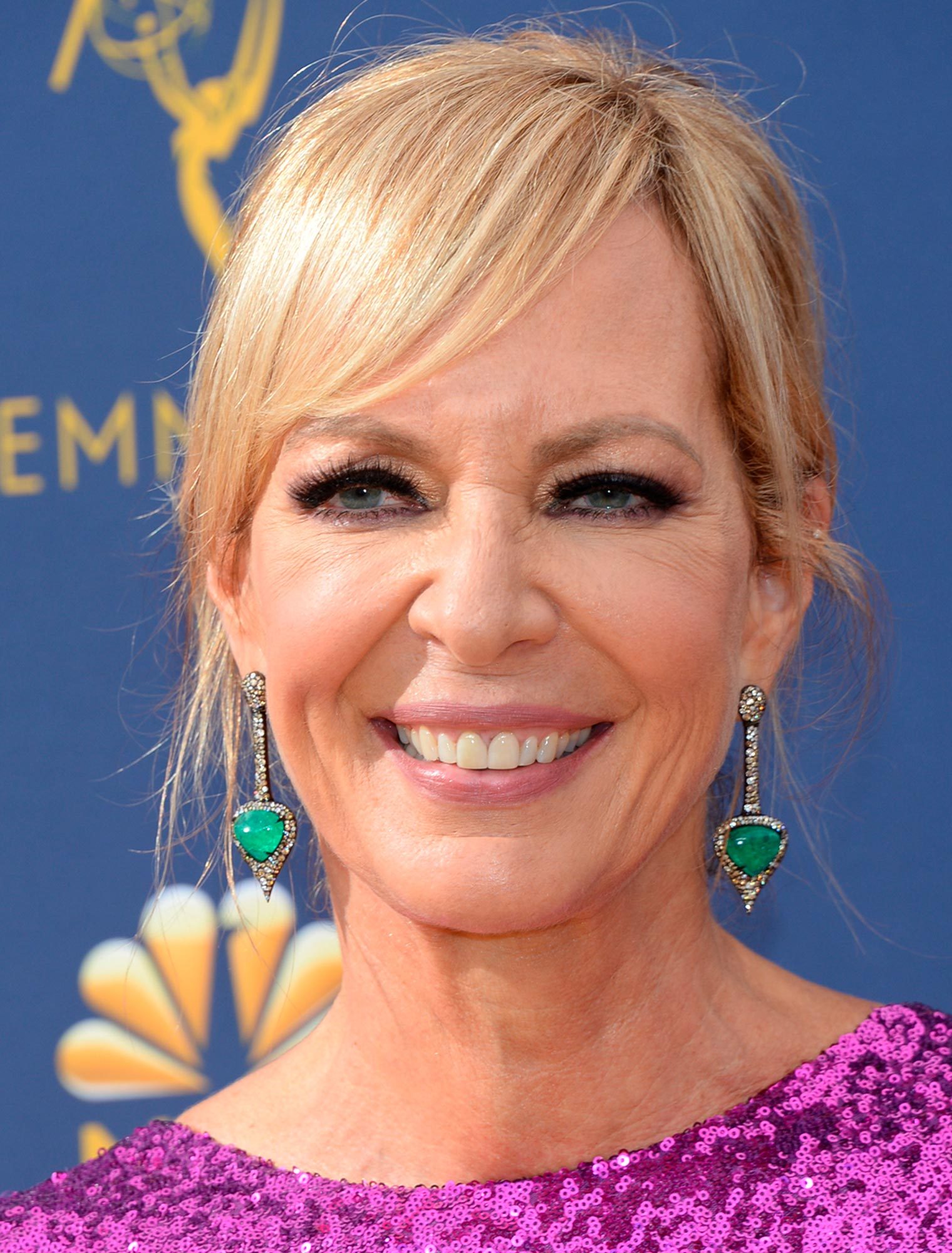 A person smiles, wearing emerald earrings and a sequined purple top, against a blue backdrop featuring the word "EMMY" and a logo.