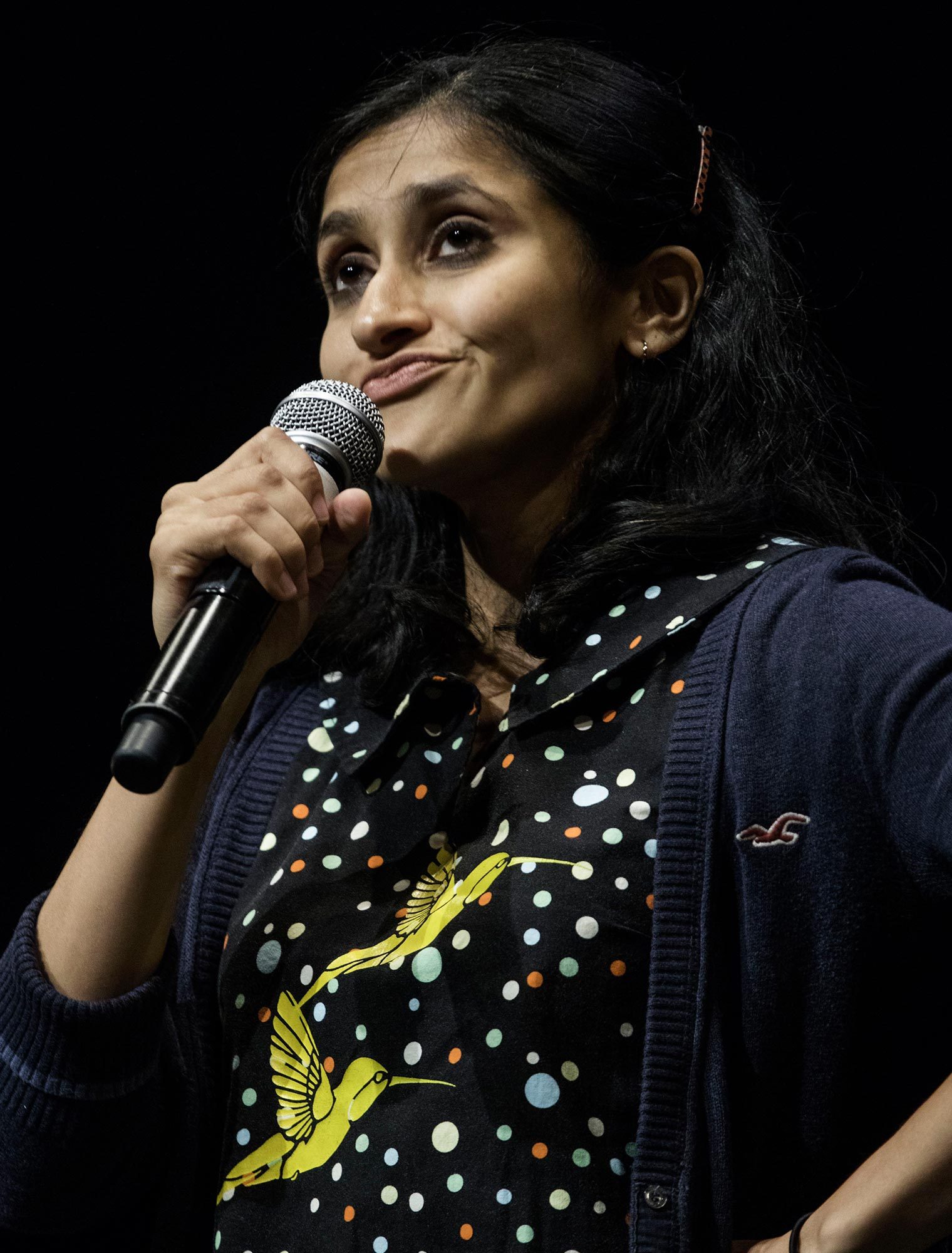 A person holding a microphone, wearing a polka dot shirt with yellow birds, stands against a black background.