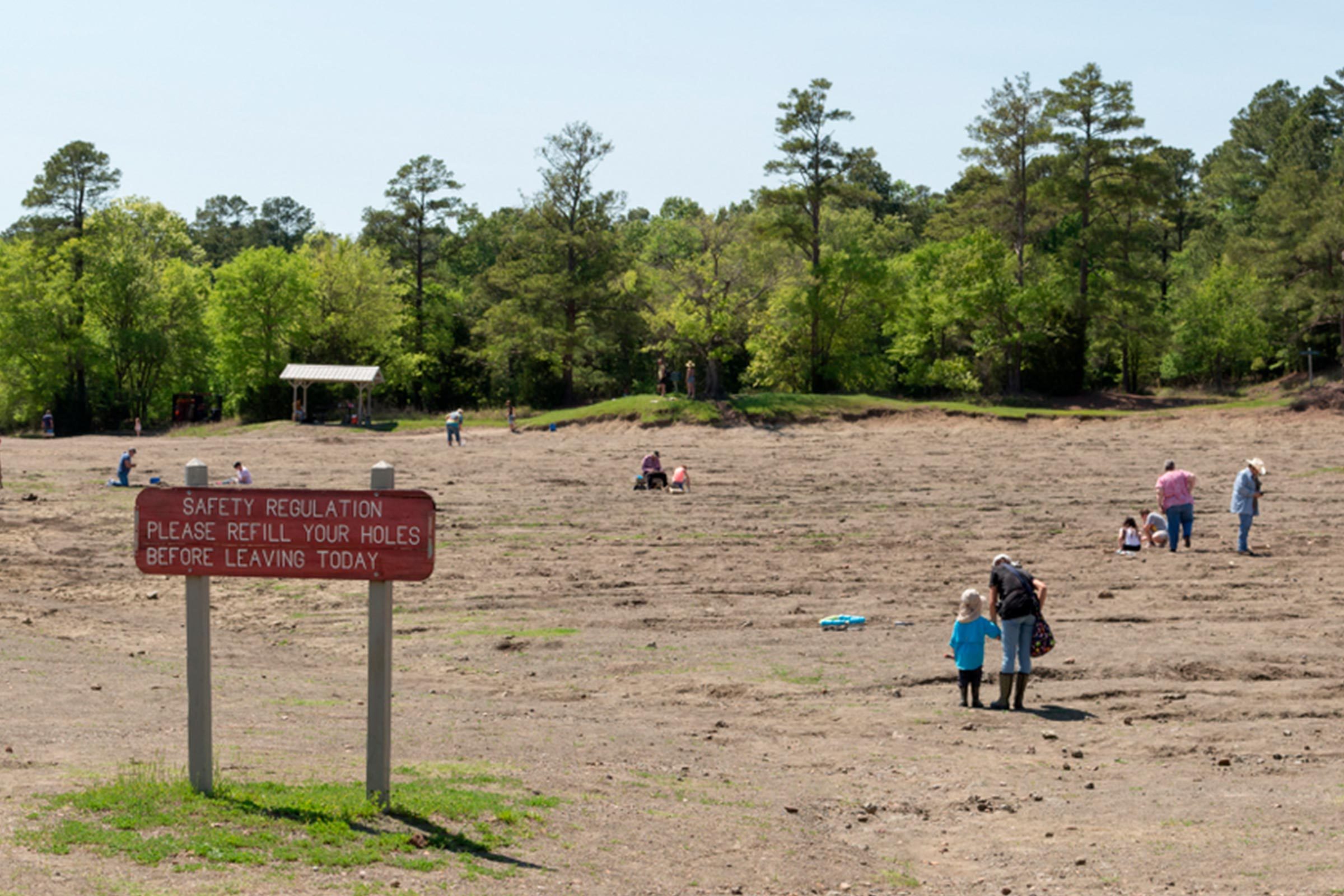 Sign warns to "refill your holes before leaving today." People dig in sandy field surrounded by trees, under a clear sky.