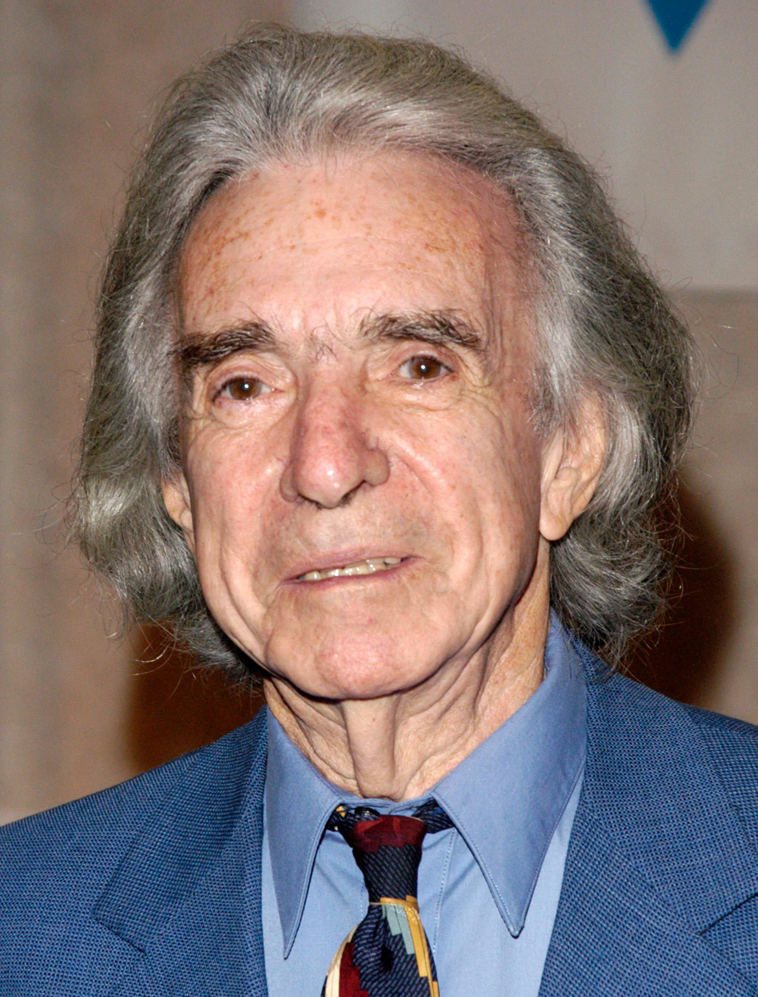 An older man with gray hair smiles slightly, wearing a blue suit and multicolored tie, standing against a neutral indoor background.