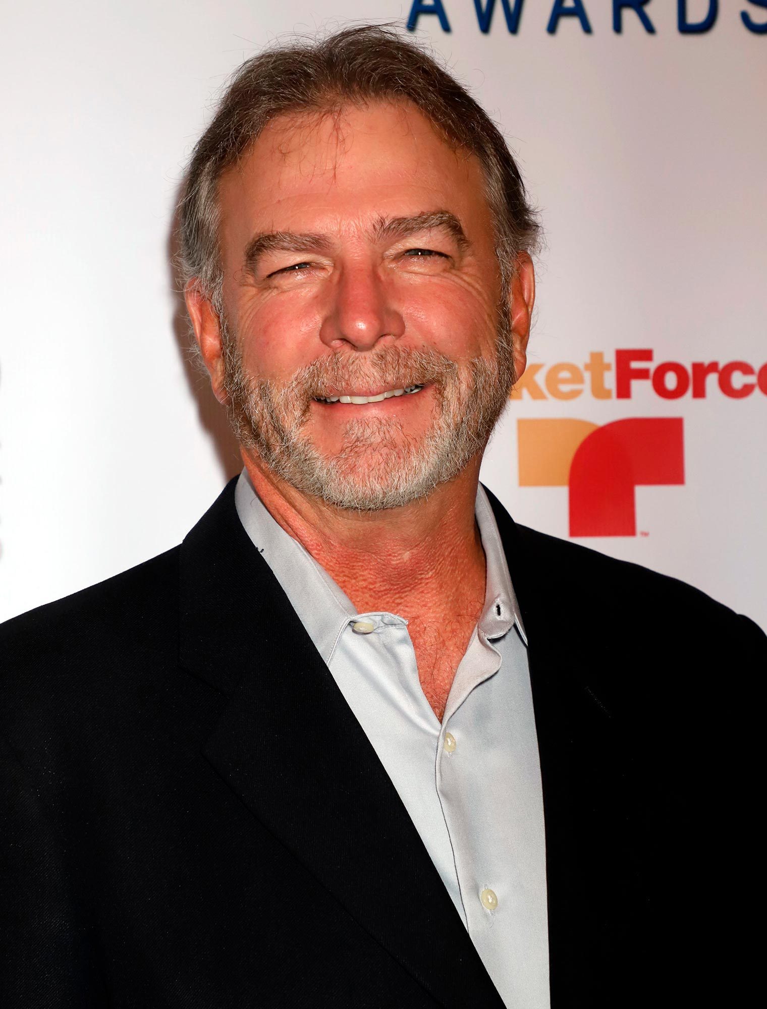 Man smiling; wears a black suit and light gray shirt; stands against a backdrop with partially visible text â€œcketForce Tâ€ with logo icon.