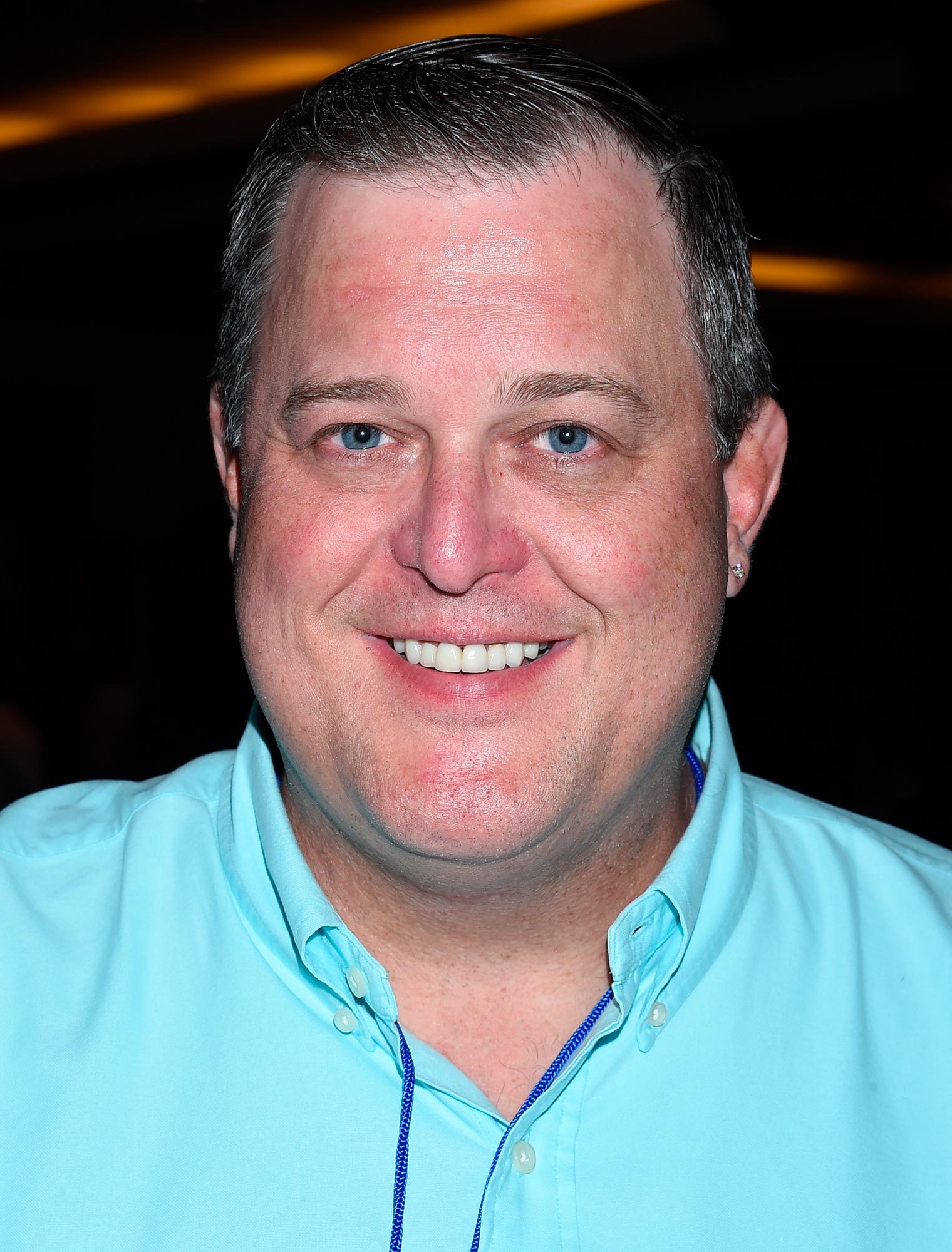 Person smiling, wearing a light blue collared shirt, standing indoors against a dark background with diffused overhead lighting.