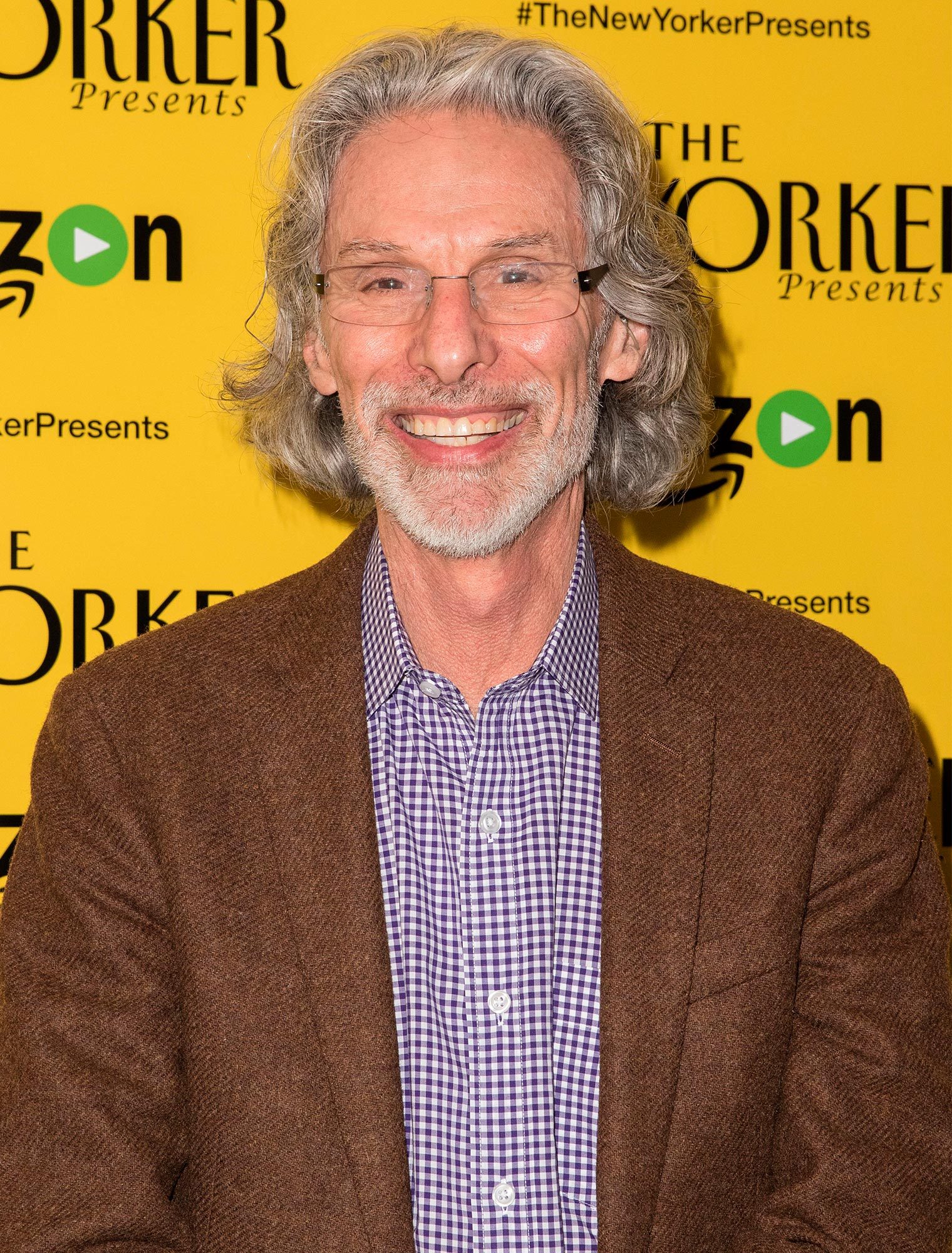 A man smiles, wearing a brown jacket and checkered shirt, with a yellow background displaying text "#TheNewYorkerPresents" and "Amazon" logo.