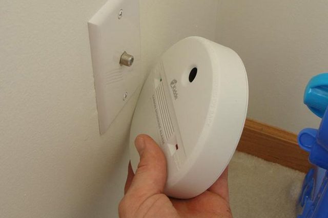 Hand holding round smoke detector near coaxial wall outlet in a carpeted corner with a plastic item nearby.