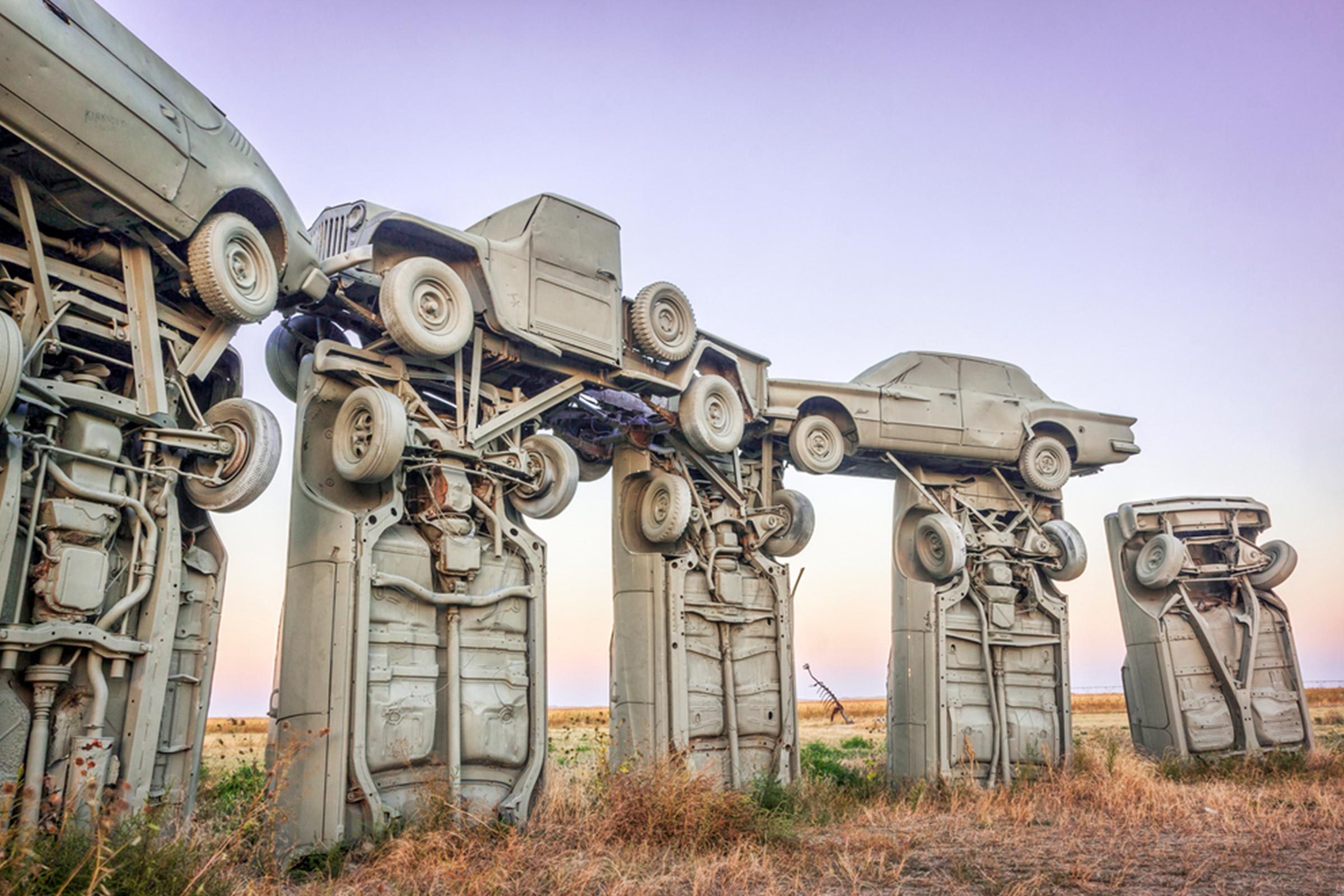 Cars are stacked vertically, resembling a stonehenge formation, set in an open grassy field with a clear sky background.