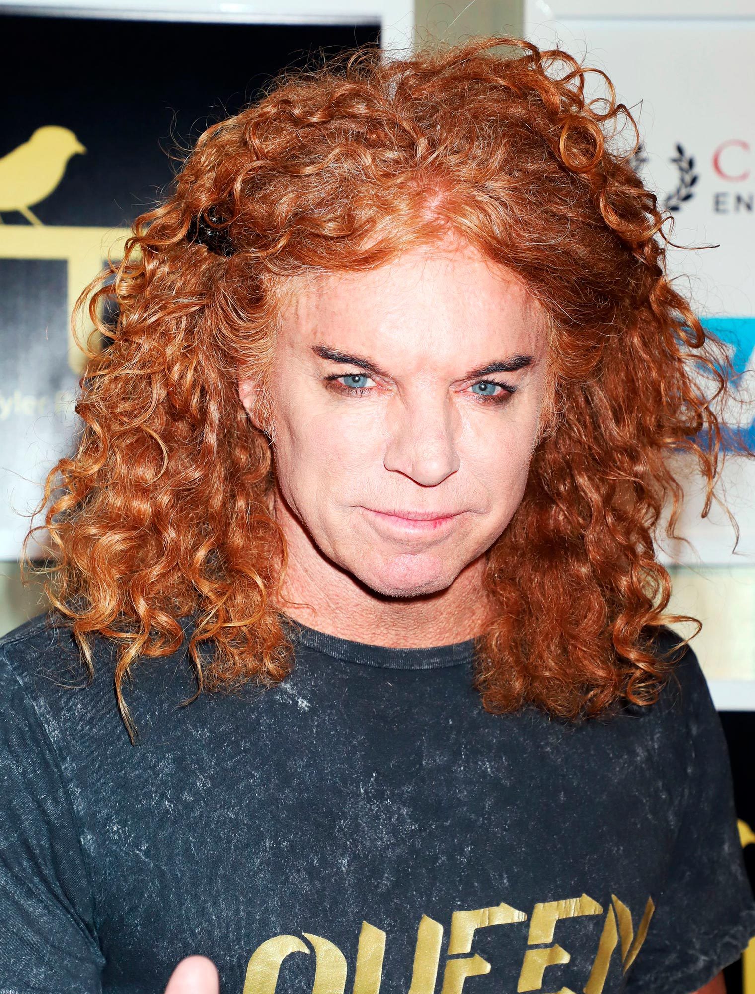 Person with curly orange hair stands casually wearing a "QUEEN" t-shirt; background includes abstract design and text, partially visible.